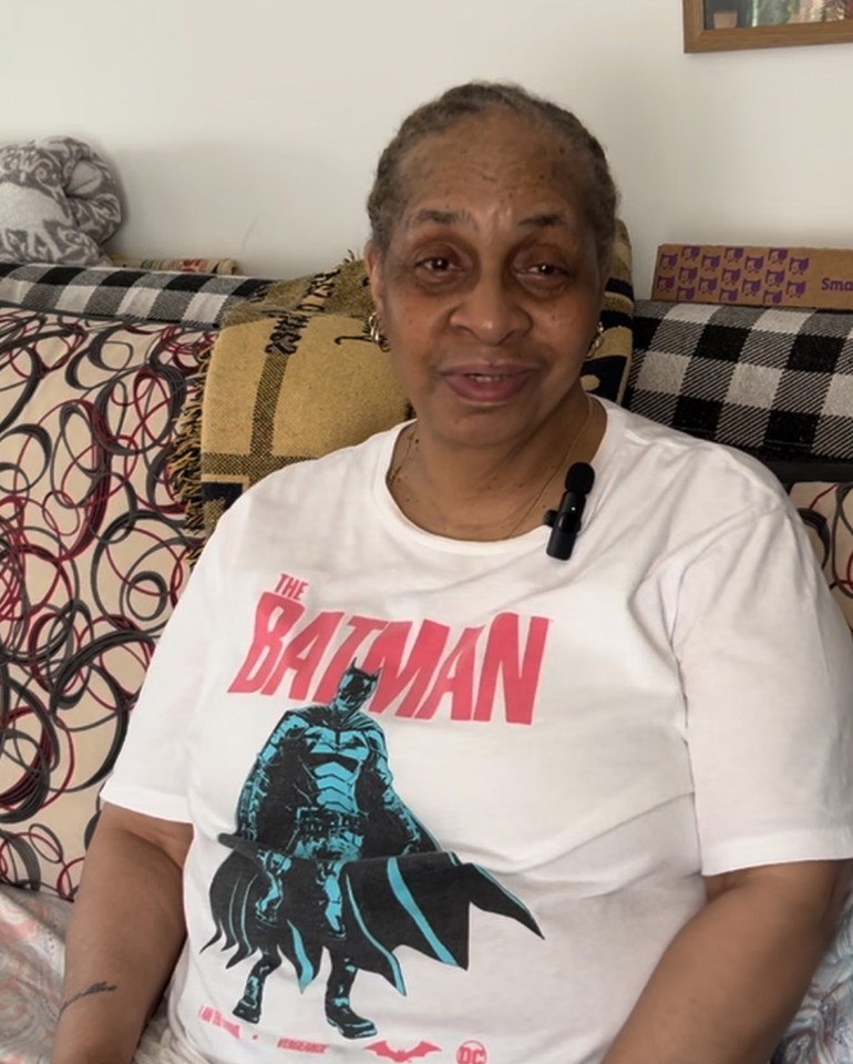 An senior woman wearing a Batman t-shirt sits on a couch and looks at the camera and smiles gently.