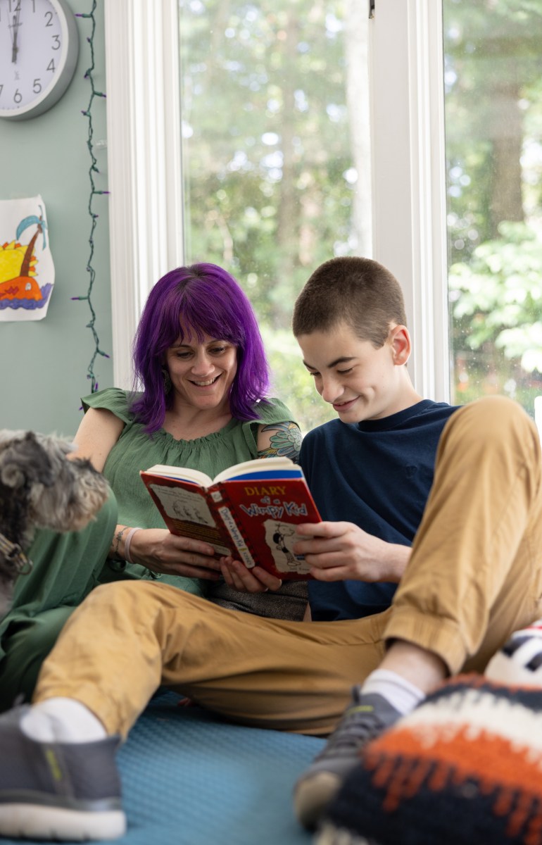 A woman with purple hair and her teenage son, who is wearing mustard colored pants, sit on a blue couch, reading a book.