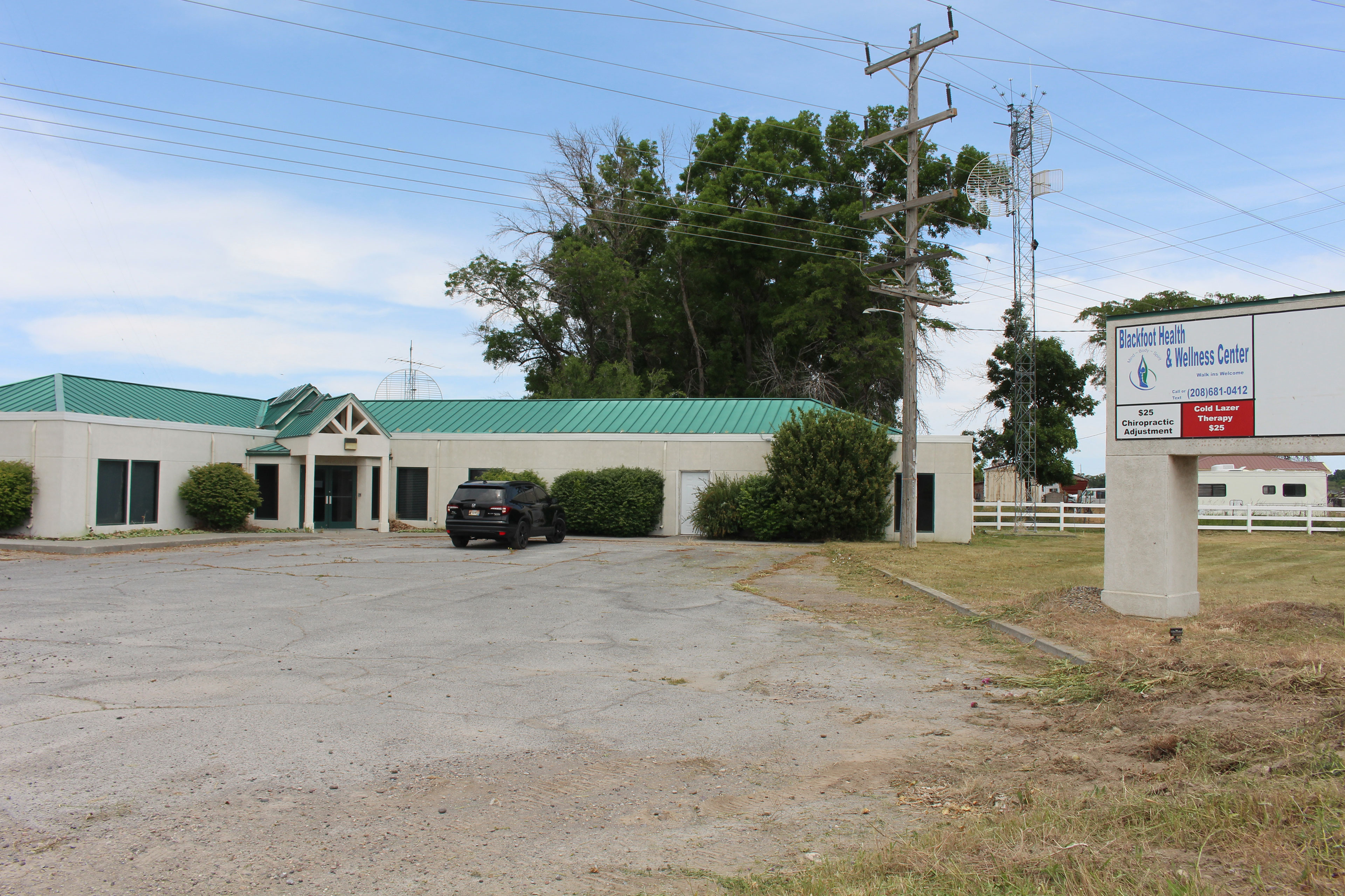 One car is parked in the small parking lot in front of a single-story beige building with a teal metal roof. A sign to the right of the building has an ad for the "Blackfoot Health & Wellness Center."