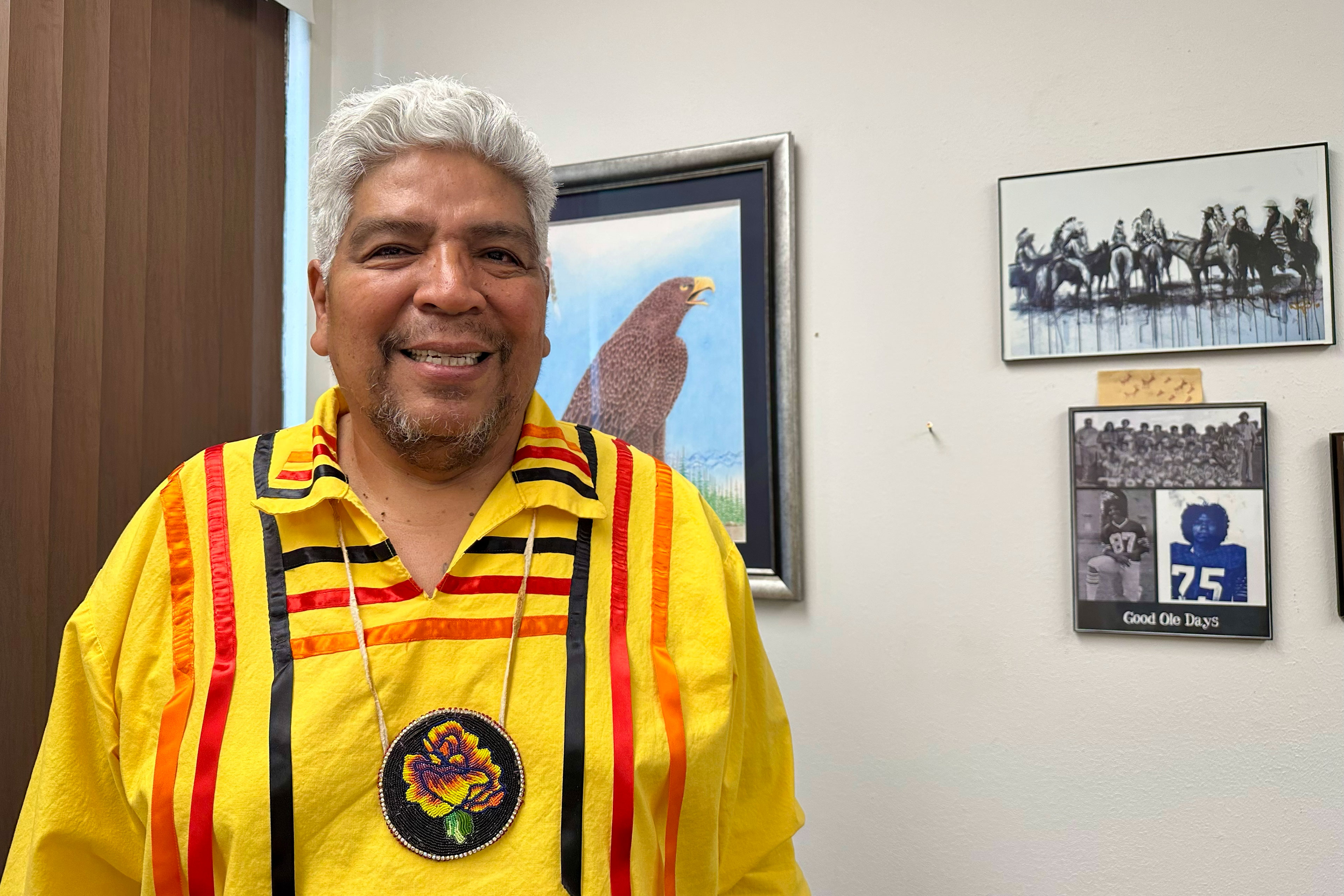 A man in a yellow shirt with orange, red, and black ribbons, and wearing a beaded necklace, stands if front of some framed pictures on a wall and smiles at the camera.