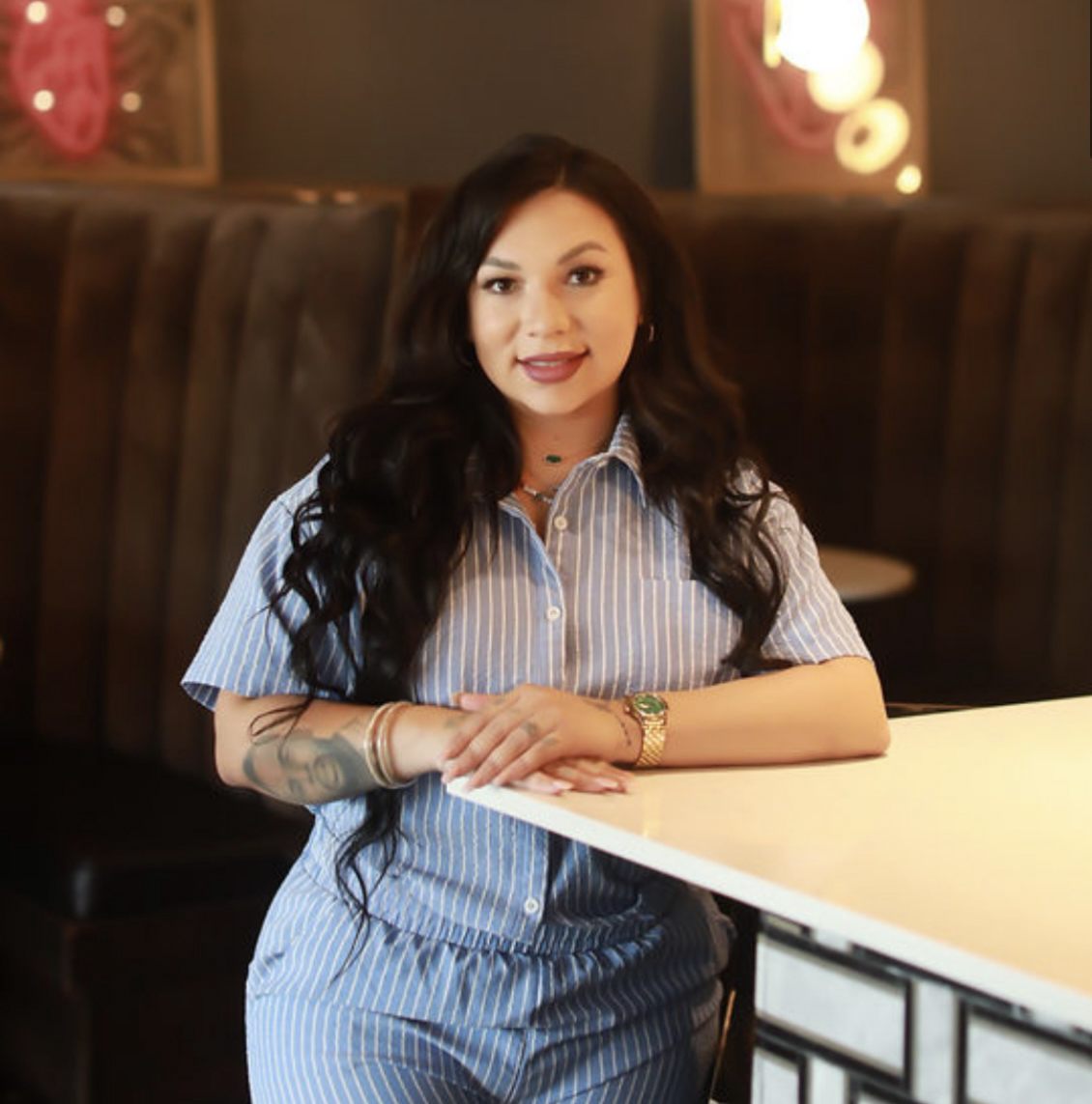 A woman with long, dark hair and a short-sleeve collared shirt stands at the corner of a bar, posing for a photo
